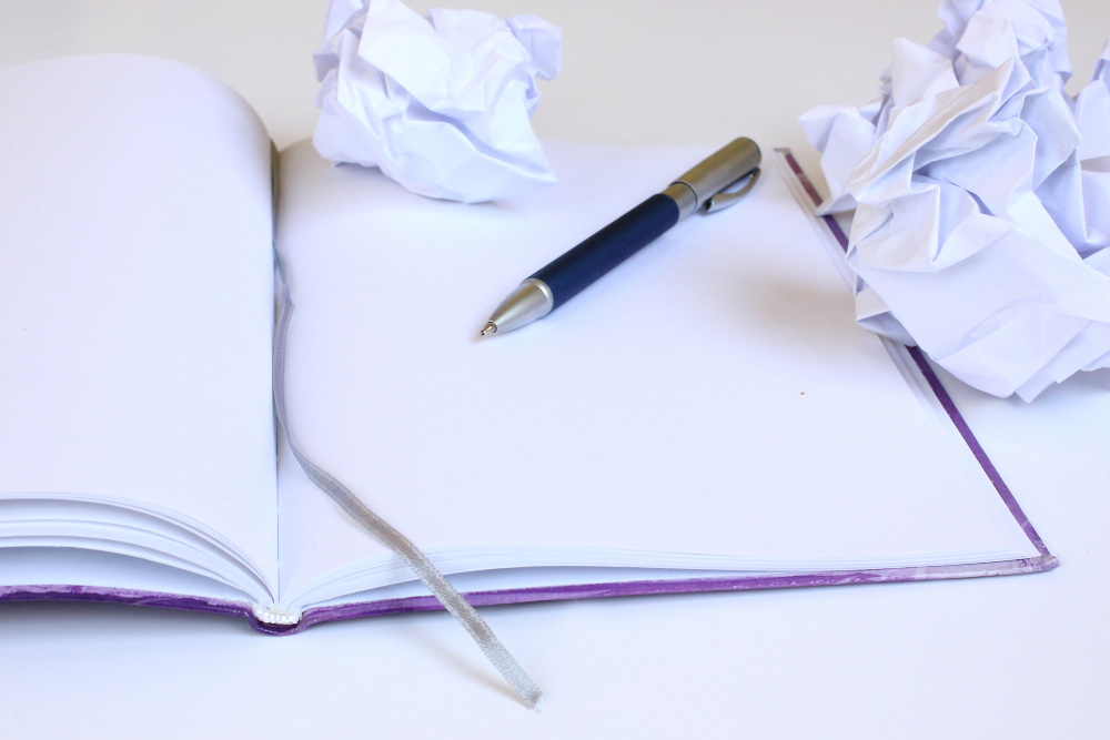Leeres Notizbuch mit Stift, daneben zusammengeknüllte Seiten symbolisch für Schreibblockaden.