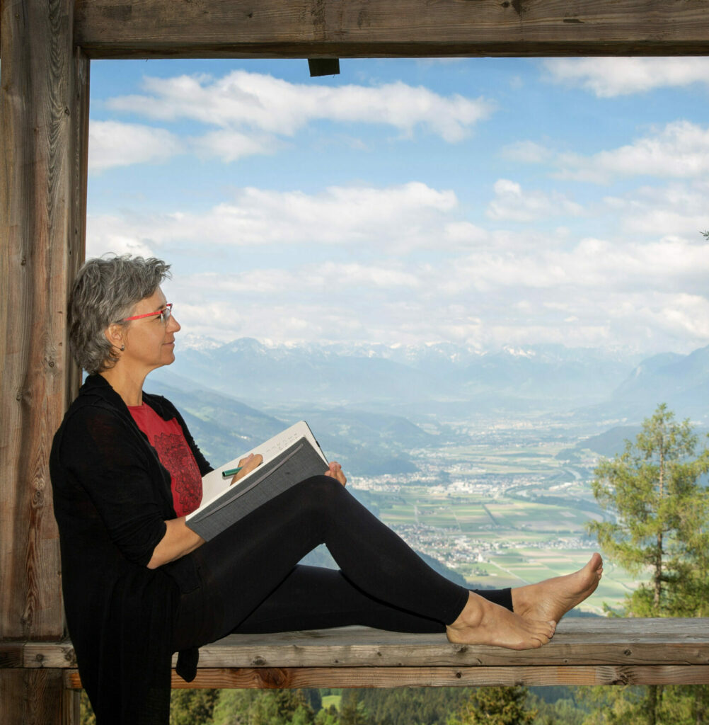 Alexandra Peischer, im Fensterrahmen sitzend und schreibend, im Hintergrund Blick ins Inntal