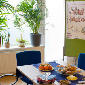 Tisch mit Brotkorb und einigen Tellern, dahinter ein Plakat "Schreibfrühstück"