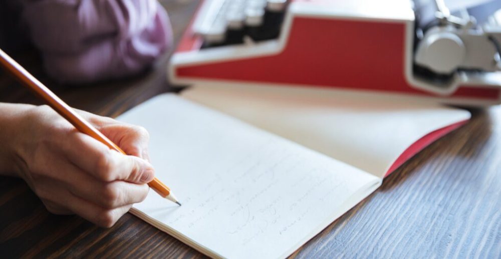 Frauenhand mit Bleistift schreibt in ein Notizbuch, daneben eine Schreibmaschine