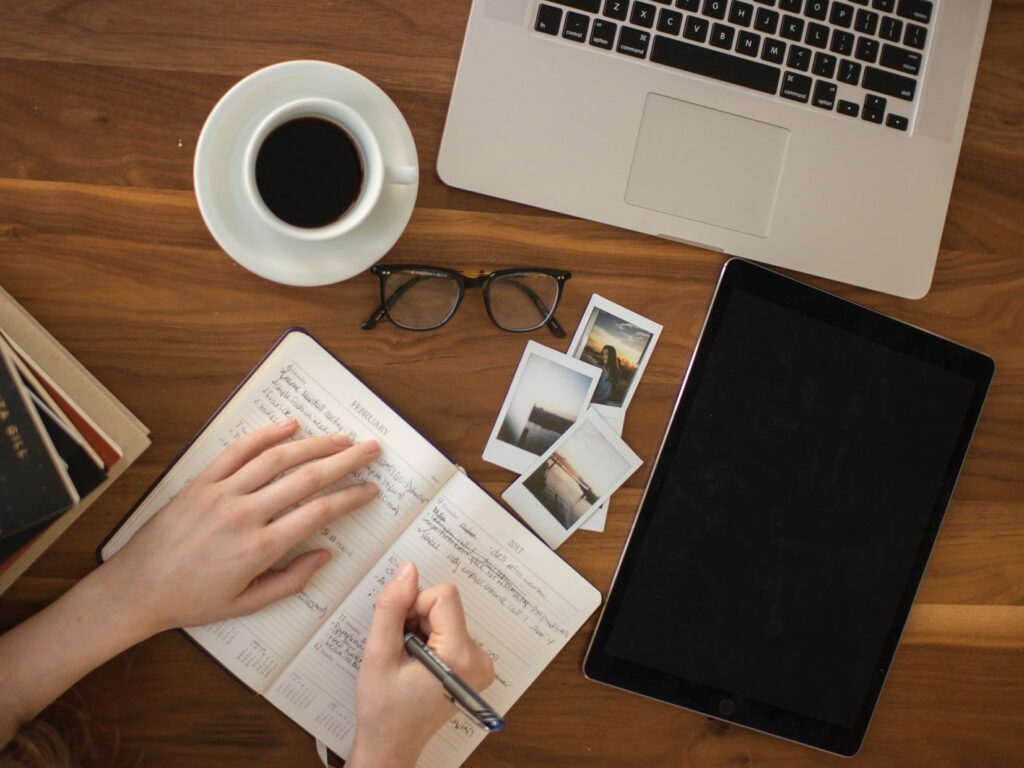 Frauenhand mit aufgeklapptem Kalender, die Notizen macht, vor ihr liegen Fotos, ein Laptop, eine Brille und eine Tasse Kaffee