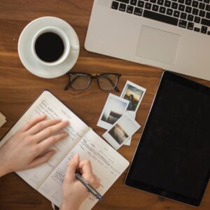 Frauenhand mit aufgeklapptem Kalender, die Notizen macht, vor ihr liegen Fotos, ein Laptop, eine Brille und eine Tasse Kaffee