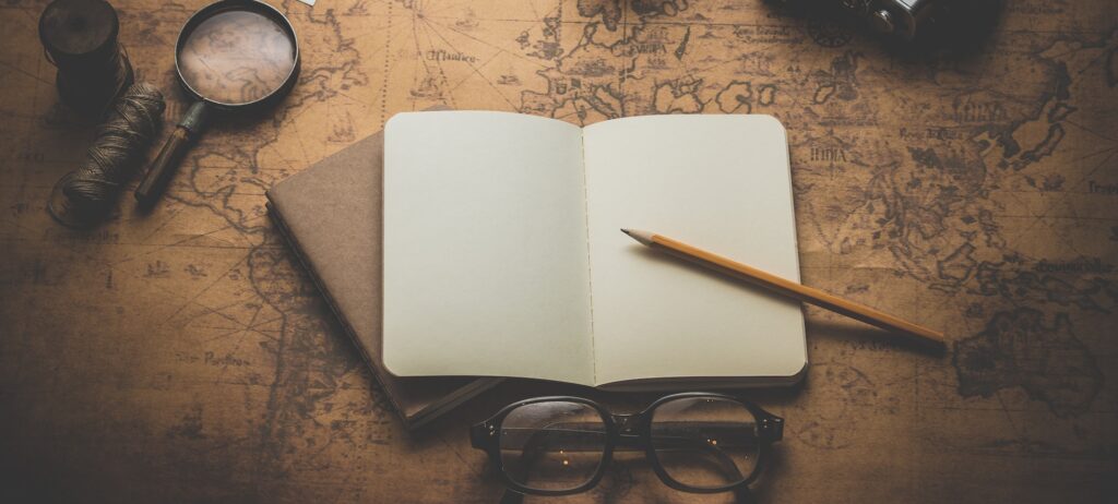 aufgeschlagenes, leeres Buch mit Bleistift auf einer alten Landkarte, daneben Brille und Lupe