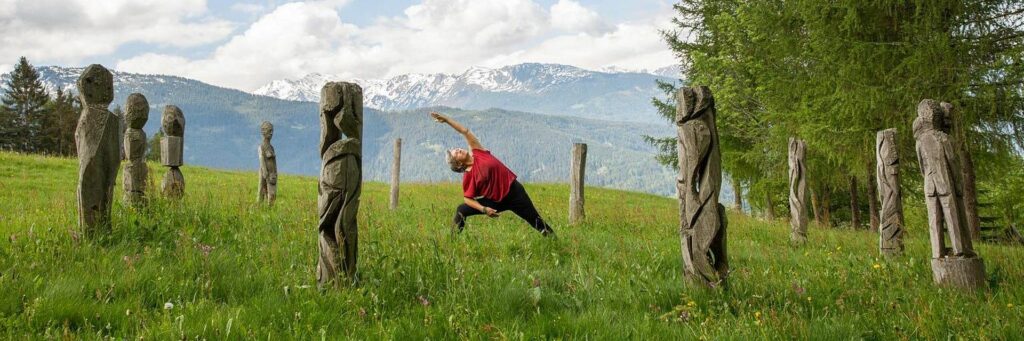 Grüne Wiese, schneebedeckte Berge im Hintergrund, eine Frau, die auf der Wiese Yoga praktiziert. Umrahmt von zwei Säulen aus Stein