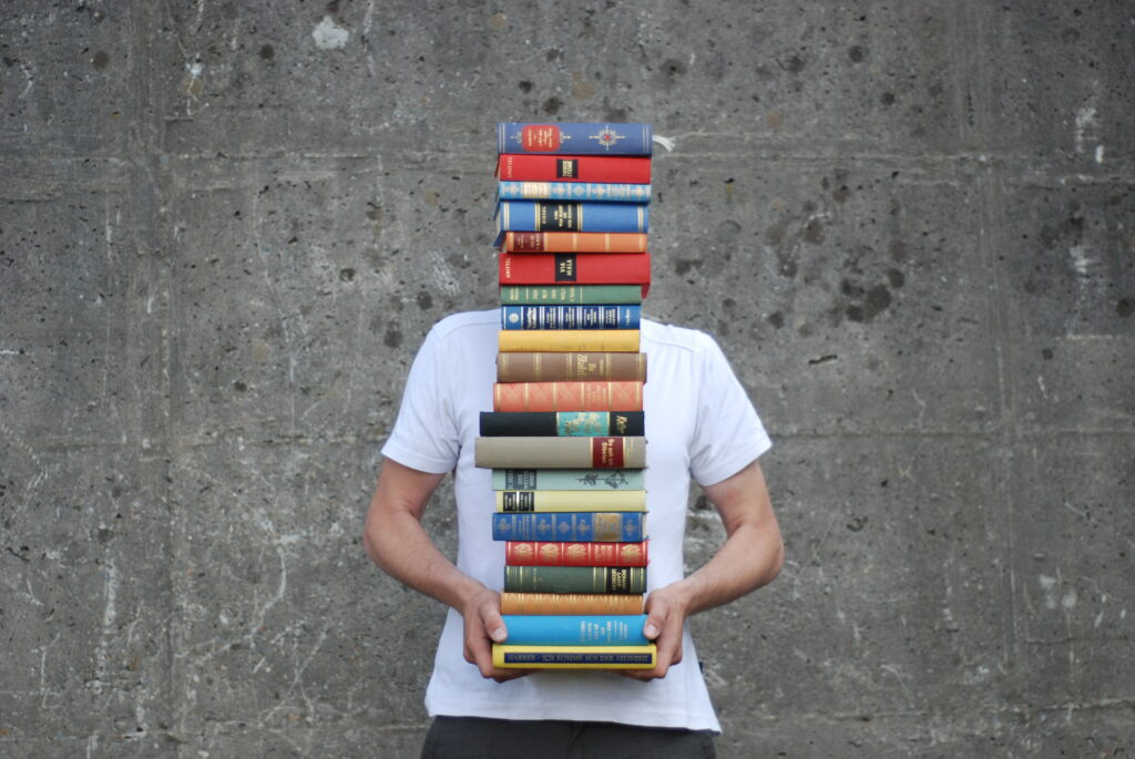 Mensch mit weissem T-Shirt hält einen Stapel bunter Bücher vor seinem Oberkörper und Kopf